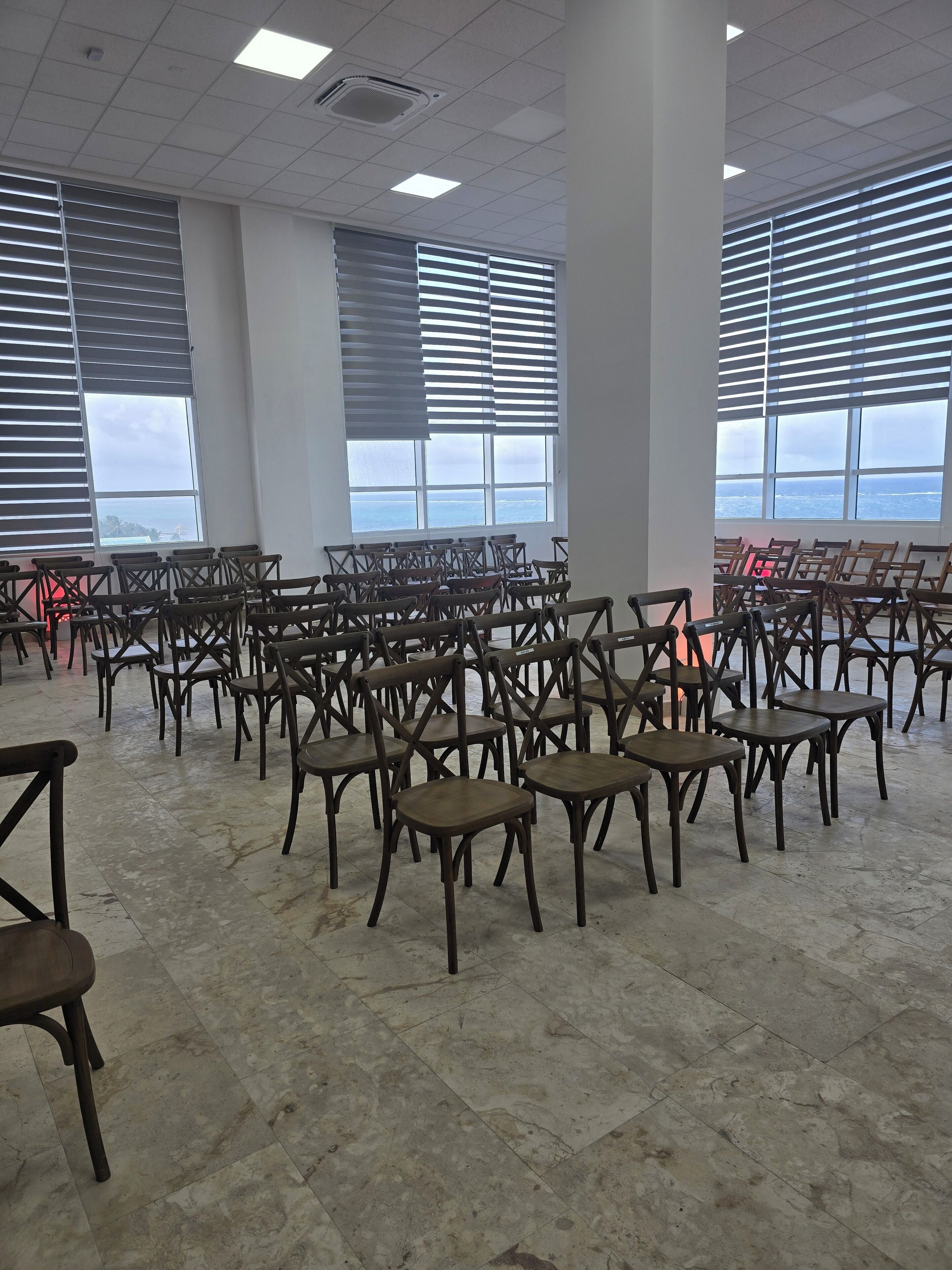 A spacious room with rows of wooden chairs and tables, large windows with blinds, and a tiled floor.