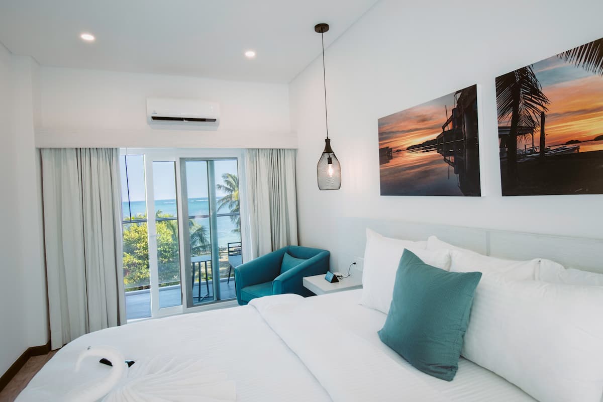 Una habitación de hotel con una cama grande, un sillón azul, una mesita de noche y vistas al océano a través de un ventanal corredero.