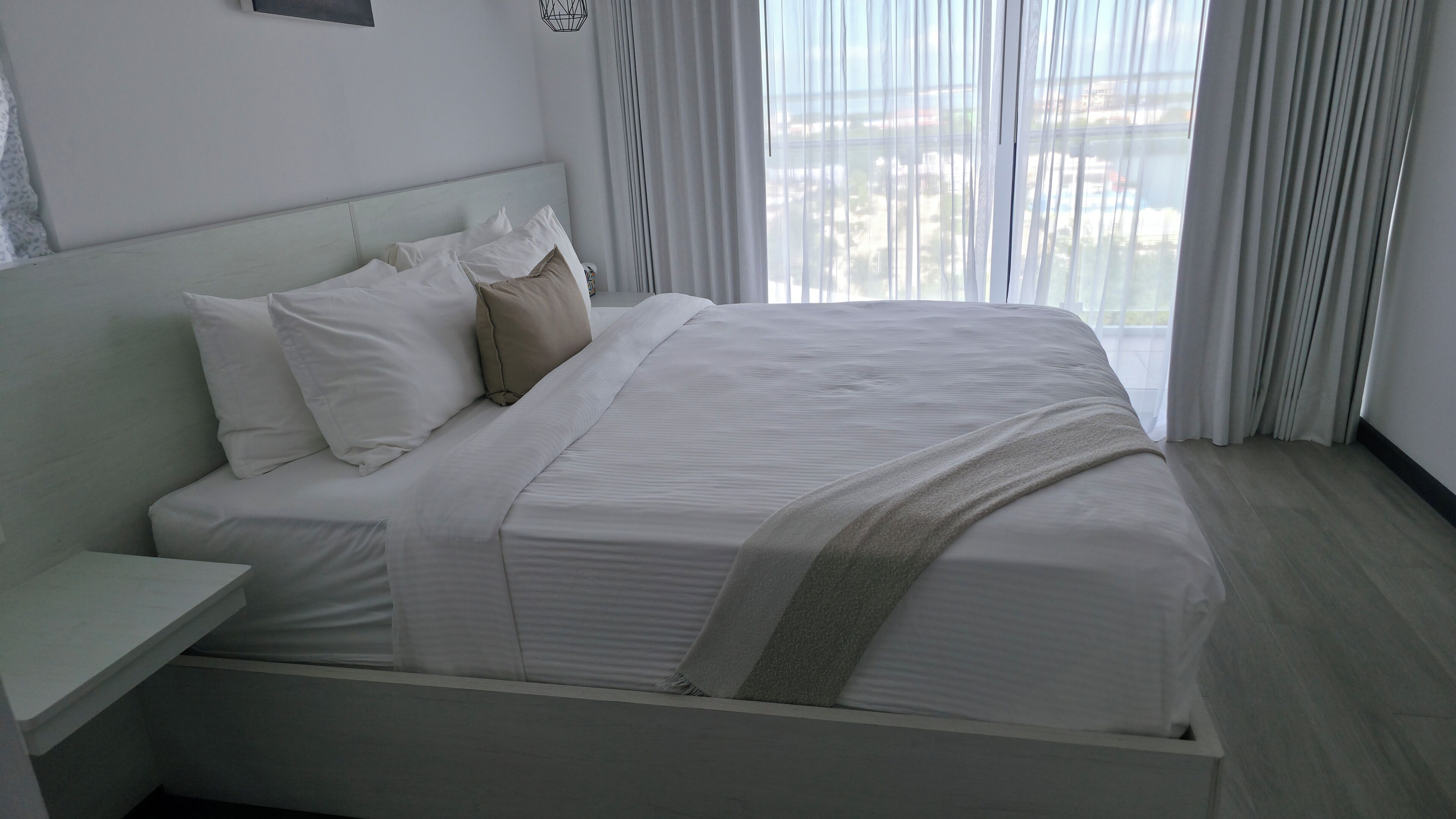 A modern hotel room with a large bed, white bedding, a bedside table, and a view of the city through sheer curtains.