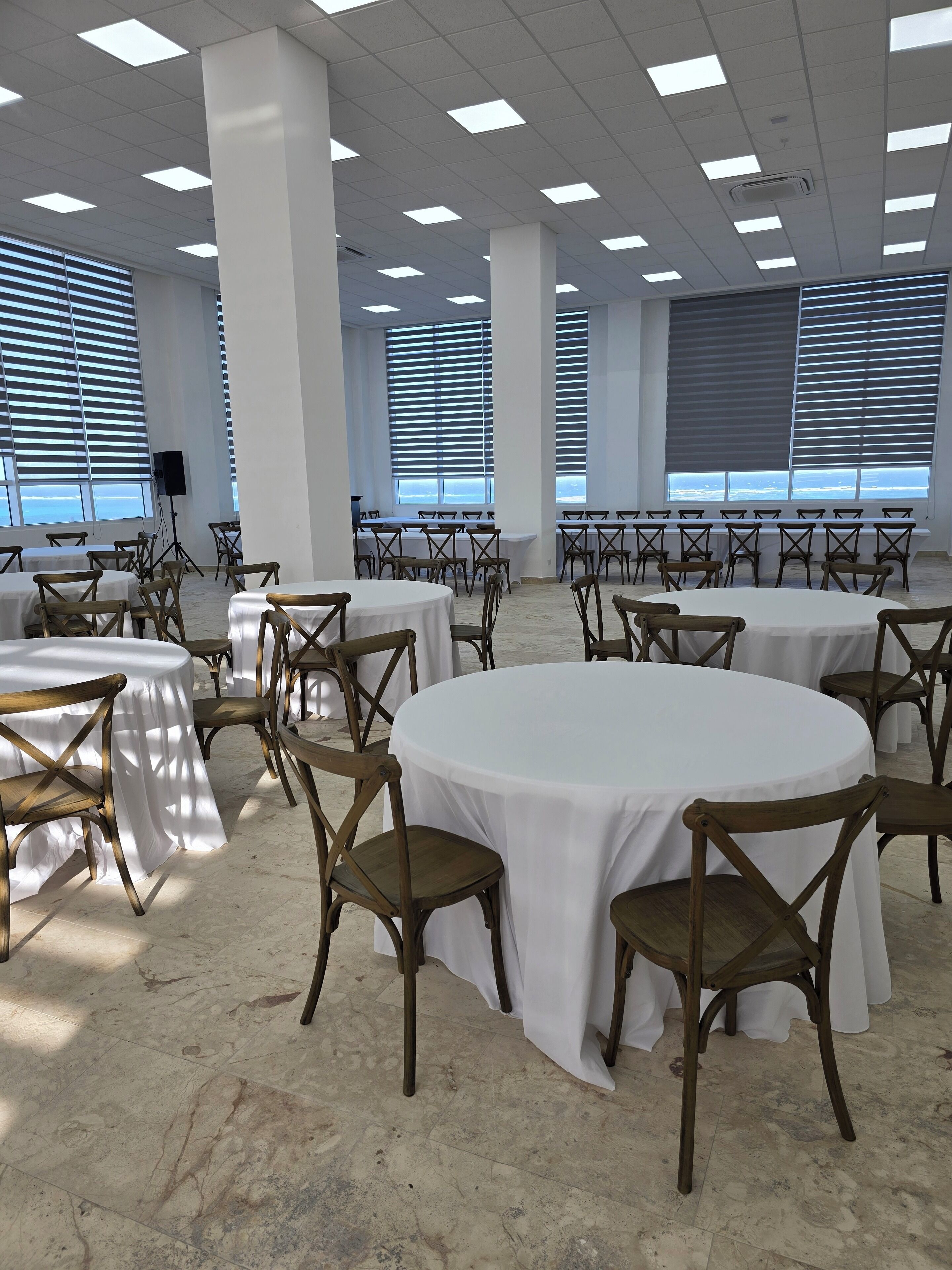 A spacious conference room with multiple round tables and chairs, large windows with blinds, and a tiled floor.