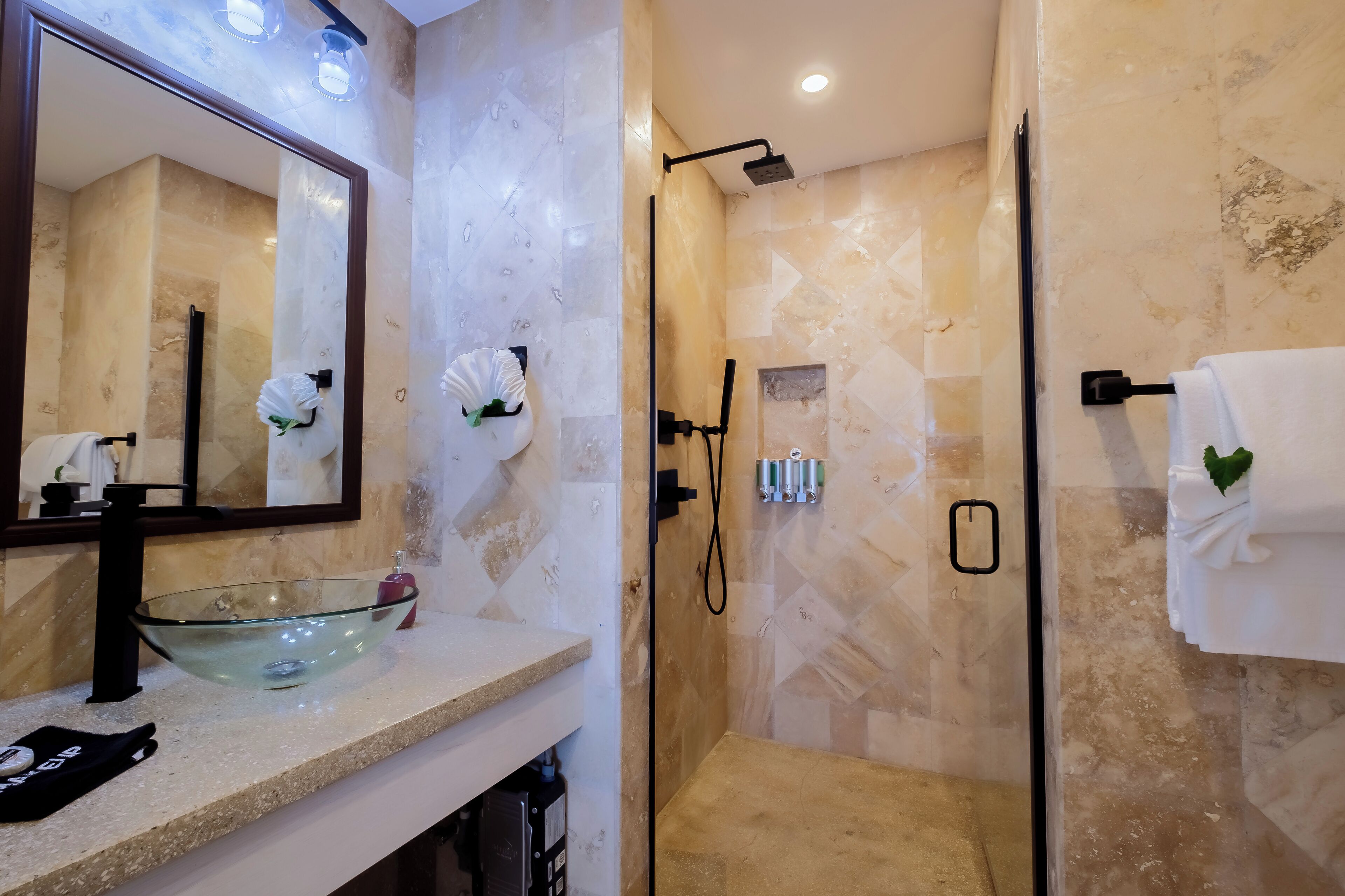 Un baño moderno con un lavabo de vidrio, un espejo grande, una ducha a ras de suelo y un perchero con una toalla blanca.