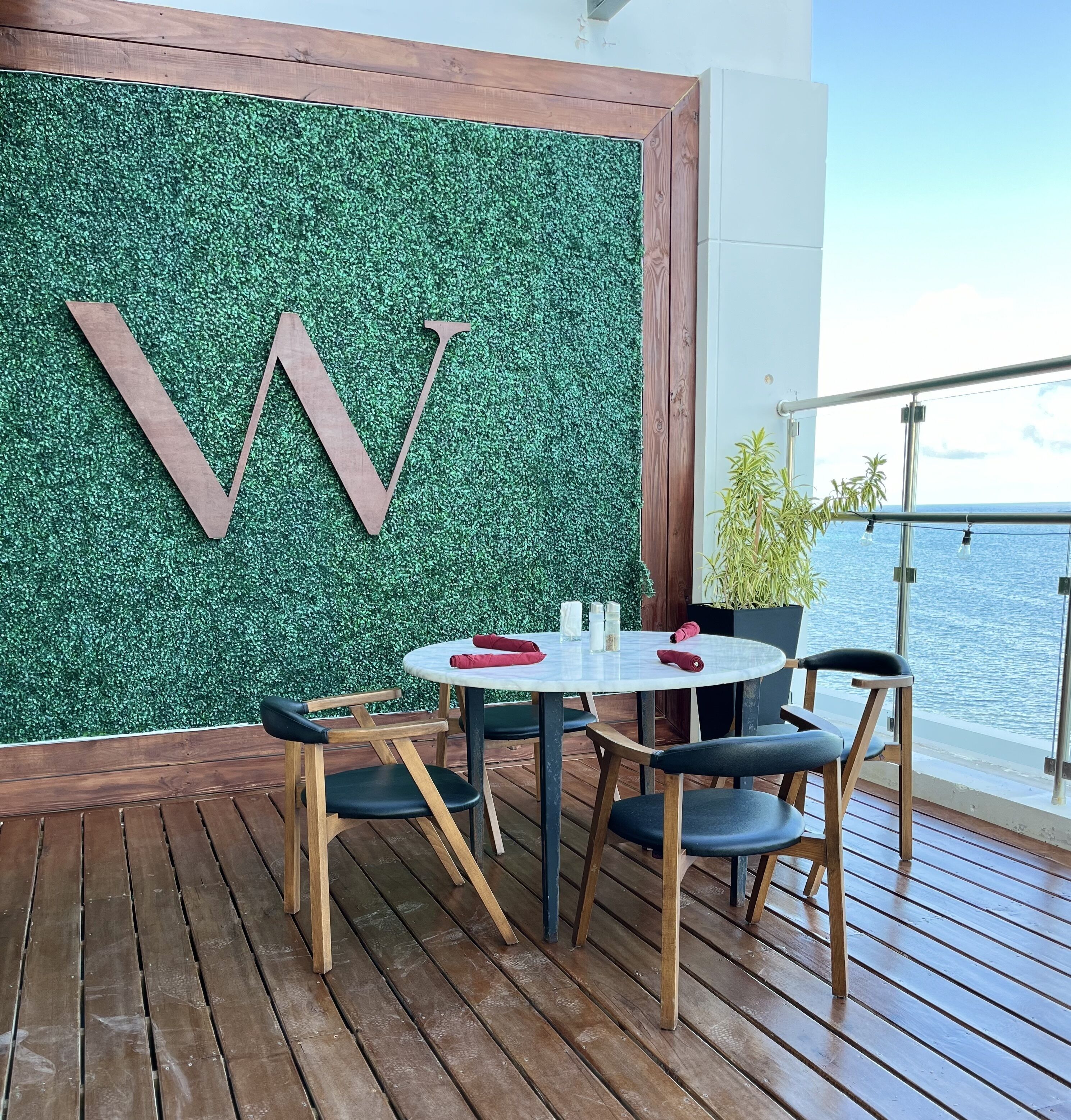 Una terraza con una mesa redonda y sillas, una pared verde con una 'W' grande, y vista al mar.