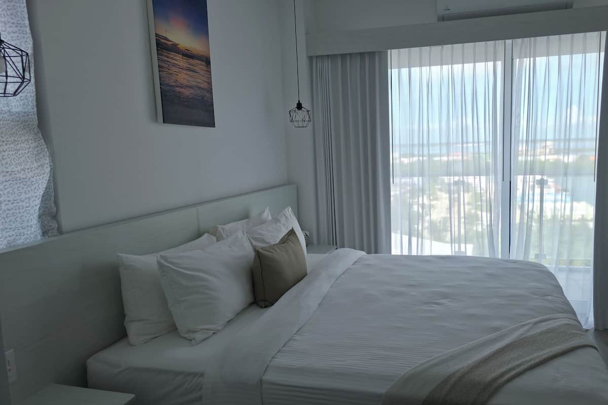 Una habitación de hotel moderna con una cama grande, ropa de cama blanca, vistas de la ciudad a través de cortinas translúcidas y una obra de arte geométrica en la pared.