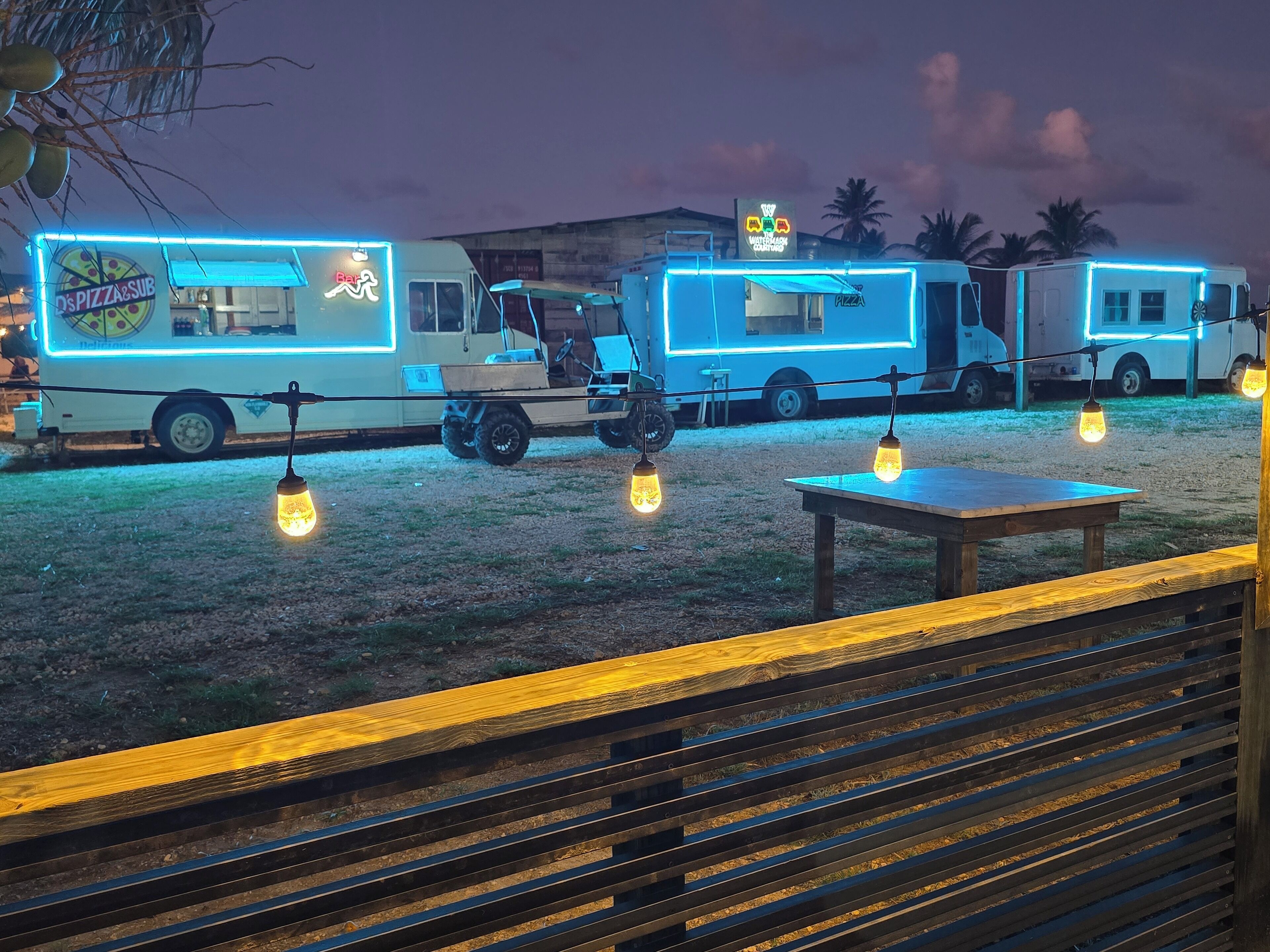A food truck area with a table and benches, and a vehicle with a neon sign.