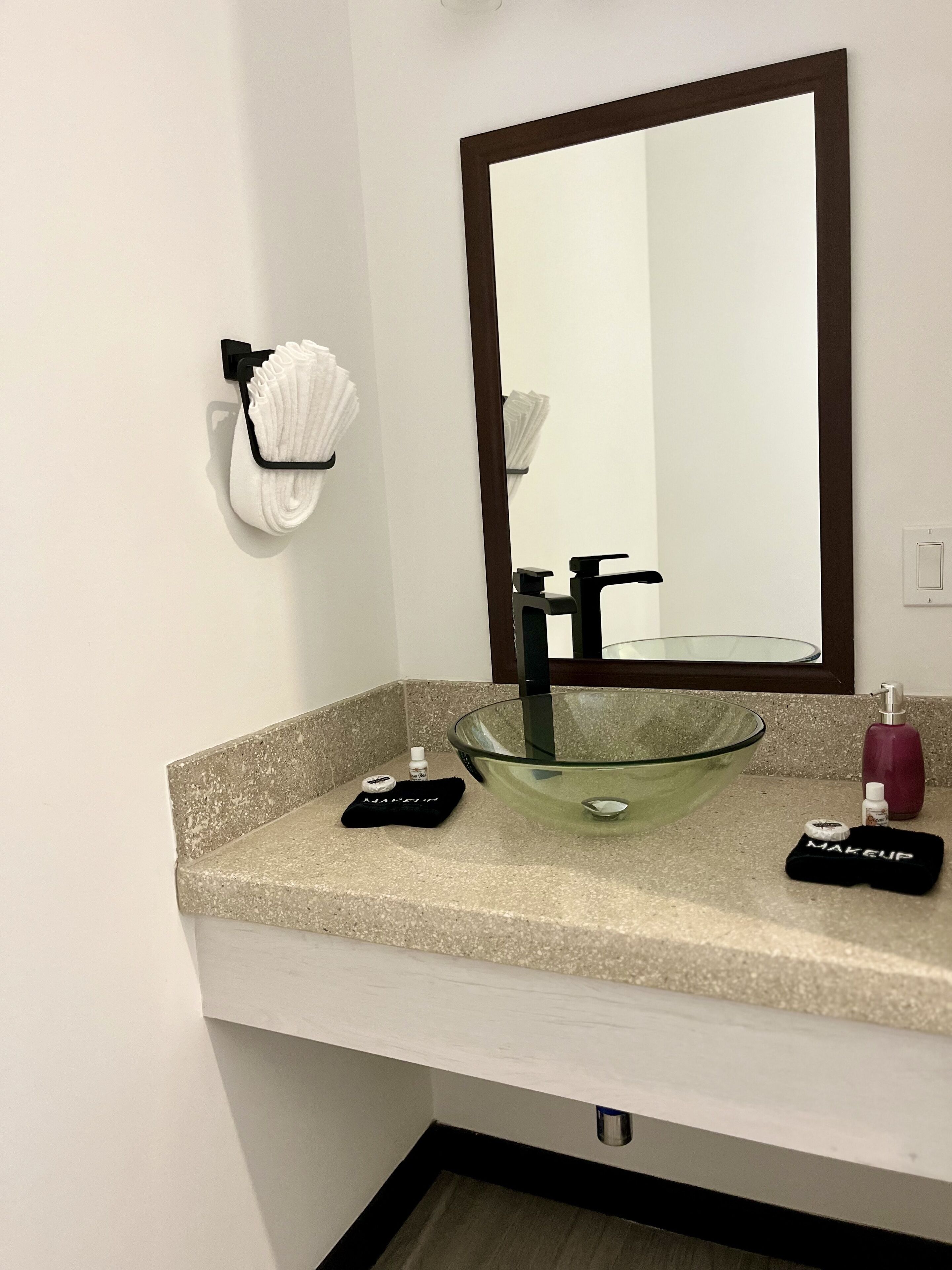Un baño con un lavabo de cristal, un grifo negro, un espejo con marco oscuro, un perchero con una toalla blanca y artículos de aseo en la encimera.