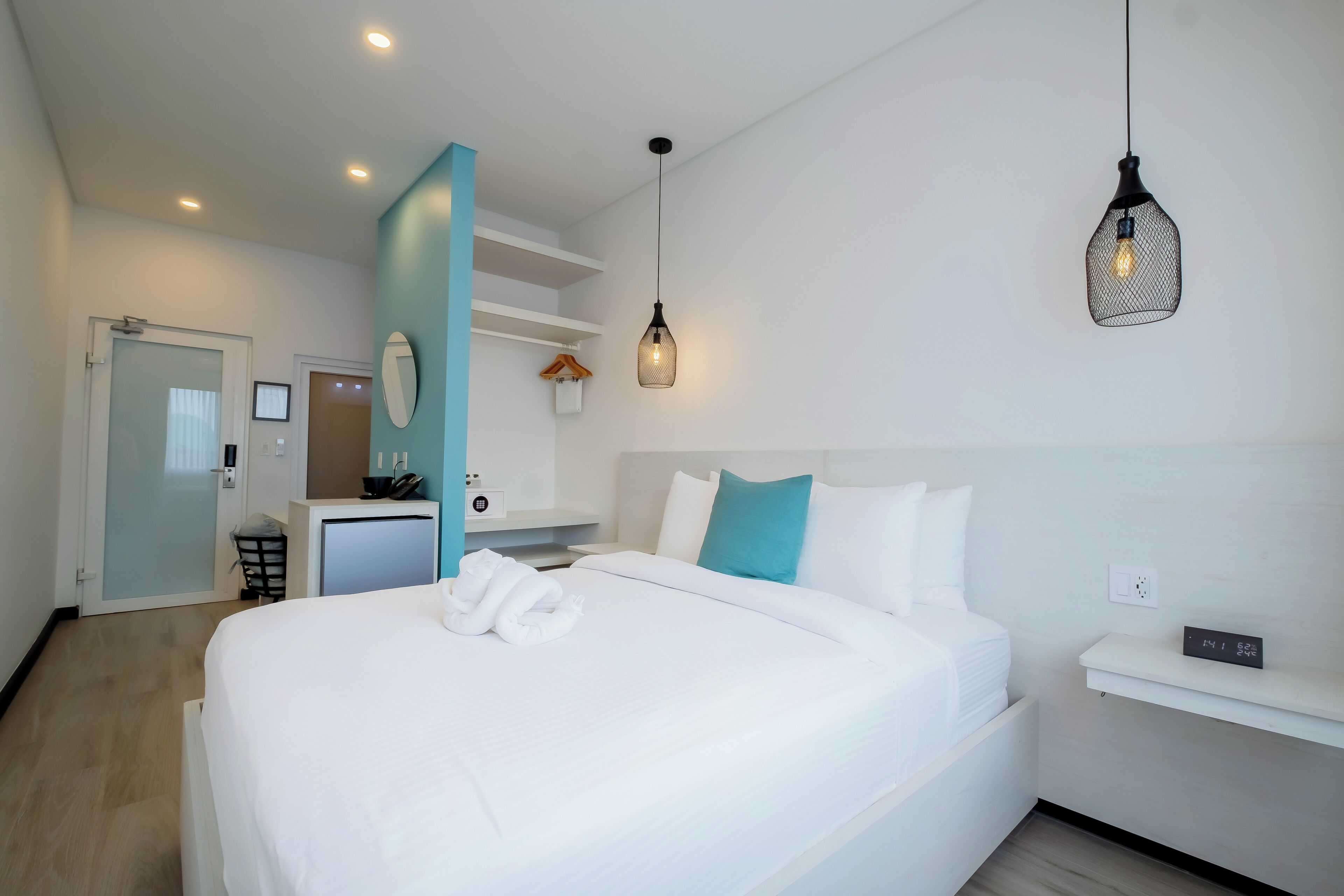 A hotel room with a neatly made bed, a small desk, a mirror, and a hanging light fixture.