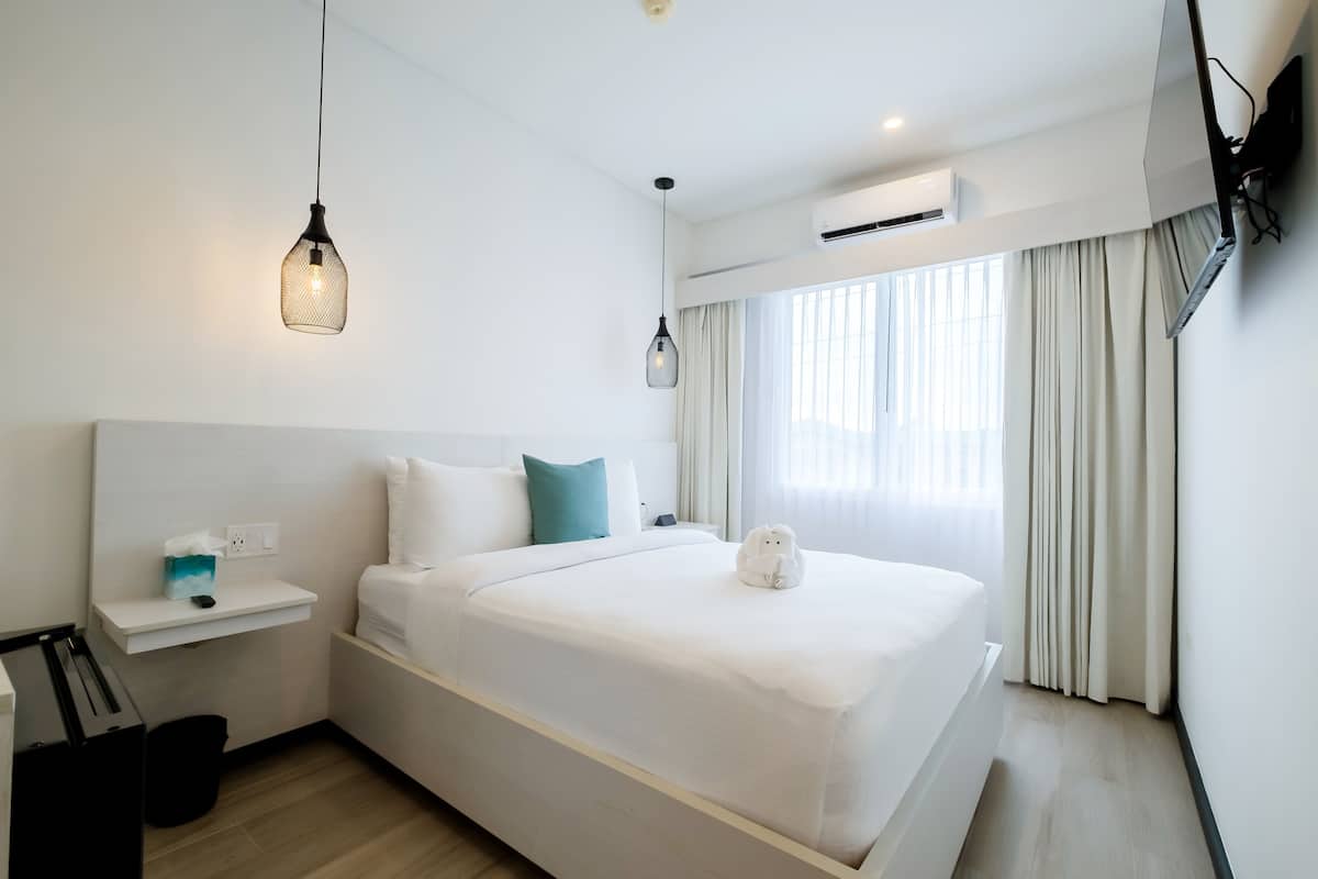 Habitación de hotel moderna con una cama bien hecha, una mesita de noche con lámpara, un televisor montado en la pared y un equipo de aire acondicionado.