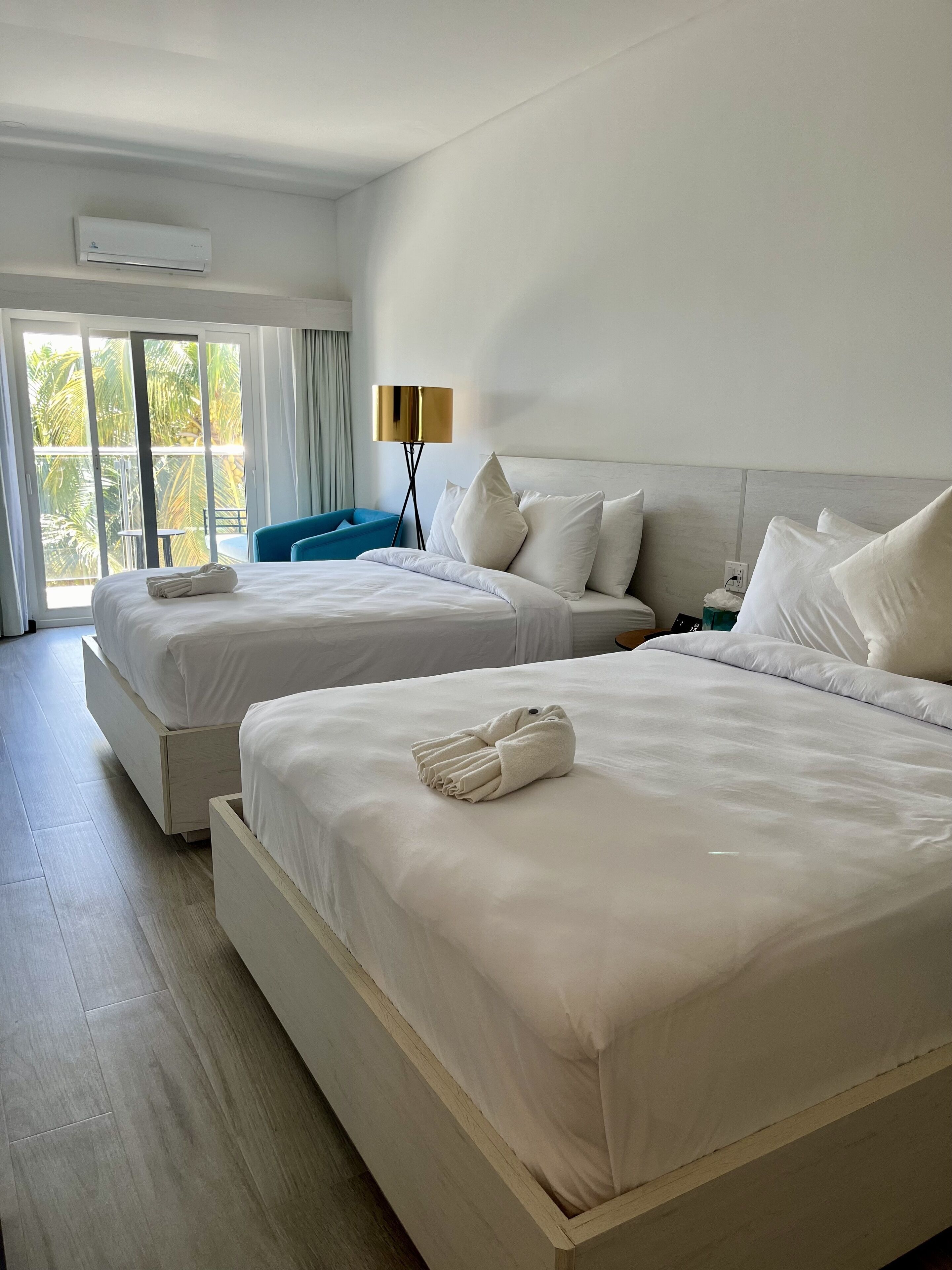 Une chambre d’hôtel avec deux lits, un balcon donnant sur des palmiers, une lampe sur pied et un climatiseur au plafond.