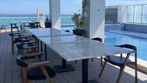 A sunlit outdoor dining area with a long table and chairs, overlooking a pool and ocean view. - The Watermark Hotel (San Pedro)