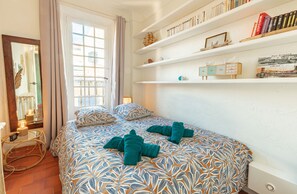 A bedroom with a bed, a mirror, a window with curtains, and a bookshelf with books and decorative items.
