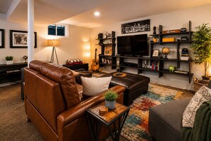 Una sala de estar con un sofá de cuero marrón, un sillón gris, una mesa de centro de madera, un televisor y una estantería.