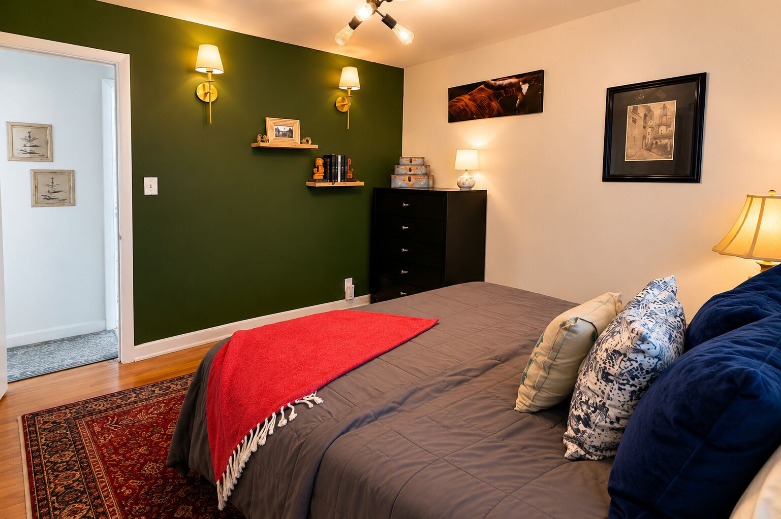 Een slaapkamer met een bed, kussens, een rode plaid, wandlampen en ingelijste foto's.