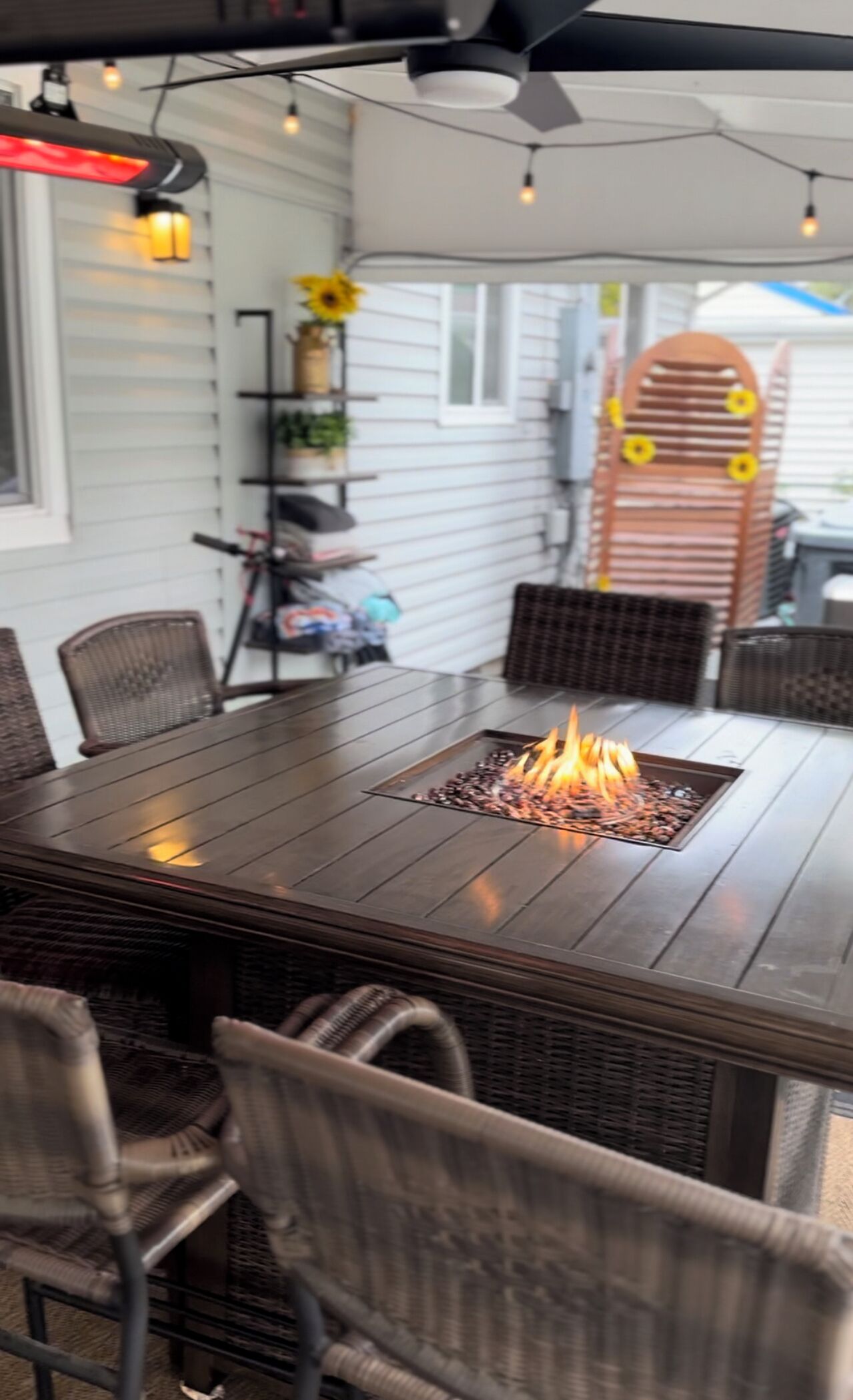 A dining area with a central fire feature, wicker chairs, and a decorative shelf with plants.