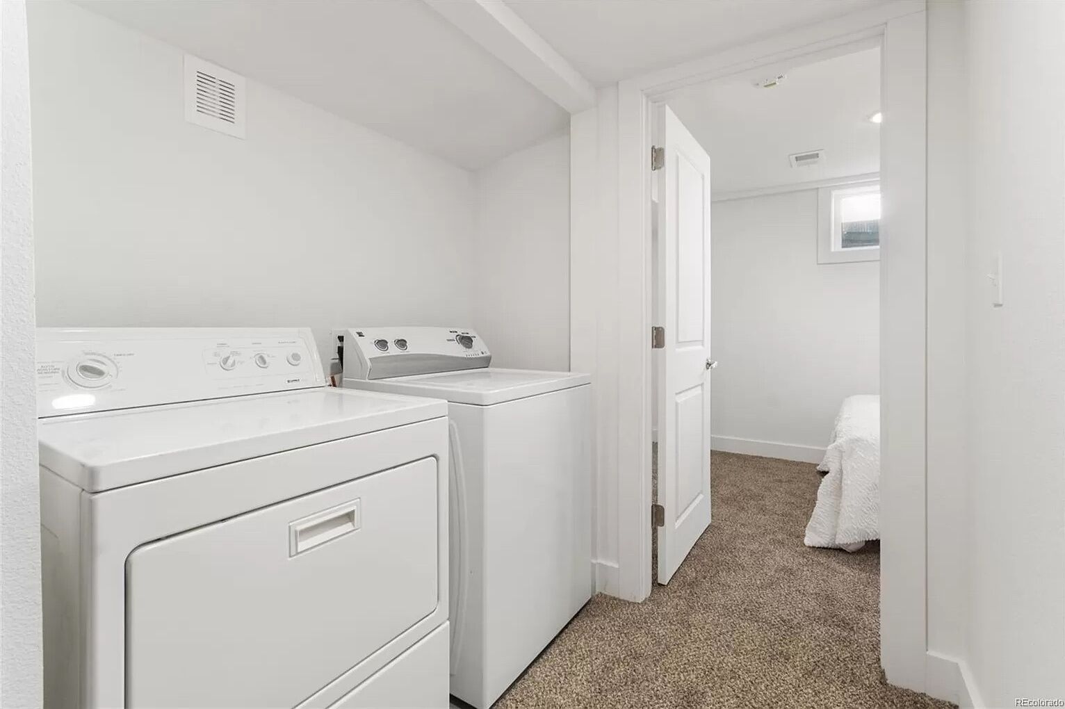 A laundry room with two white washers and dryers, a vent on the wall, and a door leading to a bedroom with a bed covered in a white blanket.