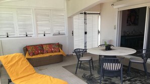 Een kamer met een gele ligstoel, een rieten bank met kleurrijke kussens, een ronde tafel met stoelen en uitzicht door een deur met een glazen ruit.