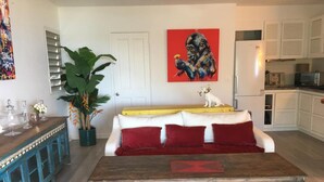 Een woonkamer met een houten salontafel, een witte bank met rode kussens, een grote groene plant en een schilderij van een gorilla aan de muur.