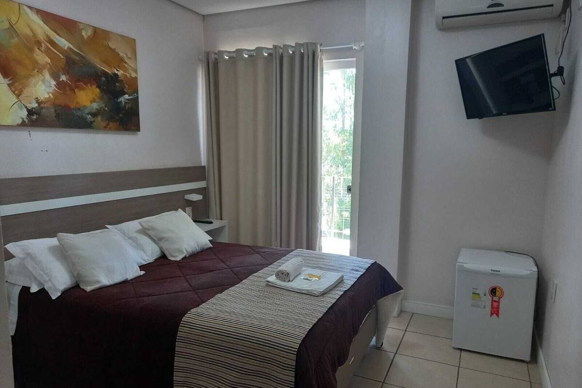 Una habitación de hotel con cama, televisión, refrigerador pequeño y ventana con cortinas.