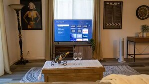 A living room with a television displaying a streaming service interface, a framed picture on the wall, and a small table with glasses and a bottle.