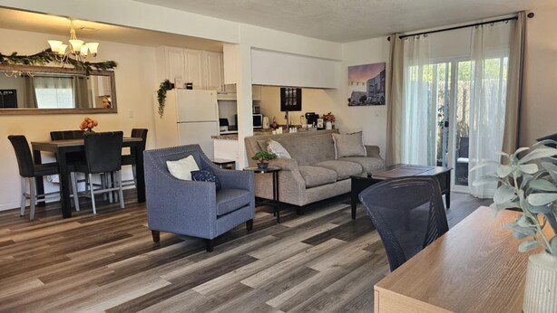 A modern living room with a sofa, armchair, dining table, and chairs. There is a kitchen area with cabinets and a refrigerator. A large window with curtains is visible.