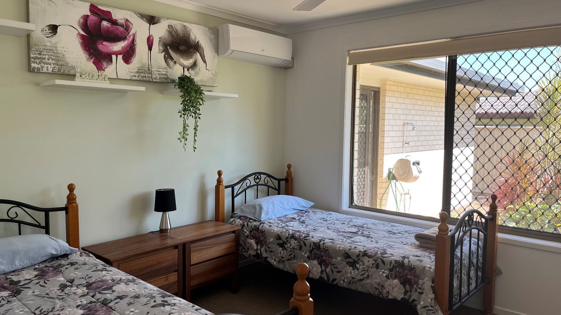 Une chambre avec deux lits, une table de chevet avec une lampe, une plante suspendue et une fenêtre donnant sur l’extérieur.