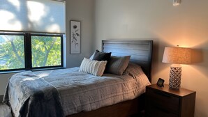 Une chambre à coucher avec un lit, une table de chevet, une lampe, un tableau encadré et une fenêtre avec des stores.