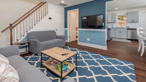 A modern living room with a gray sofa, a coffee table with books, a wooden staircase, and a kitchen in the background. - Beautiful Renovated Townhome w/parking (Holden)