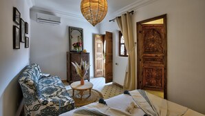 Una habitación con un sofá estampado, una mesa redonda con un florero, una cama con una colcha y una puerta grande de madera.