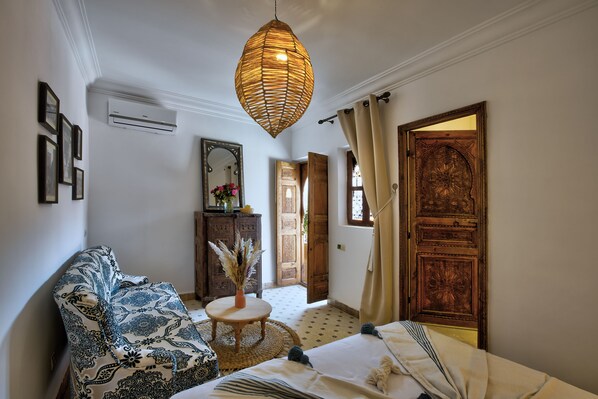 A room with a patterned sofa, a round table with a vase, a bed with a blanket, and a large wooden door. - Riad Arabkech (Marrakech)