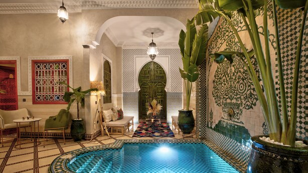 Una piscina interior lujosa con elaborados trabajos en azulejos, una zona de estar con un tapete estampado y una planta decorativa grande.