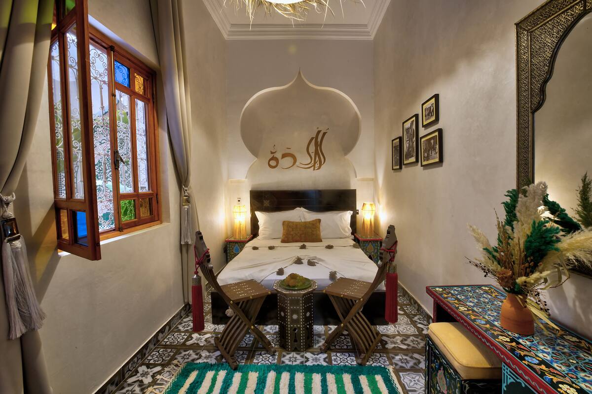 A traditional room with a bed, a small table, chairs, a decorative mirror, and a patterned rug.