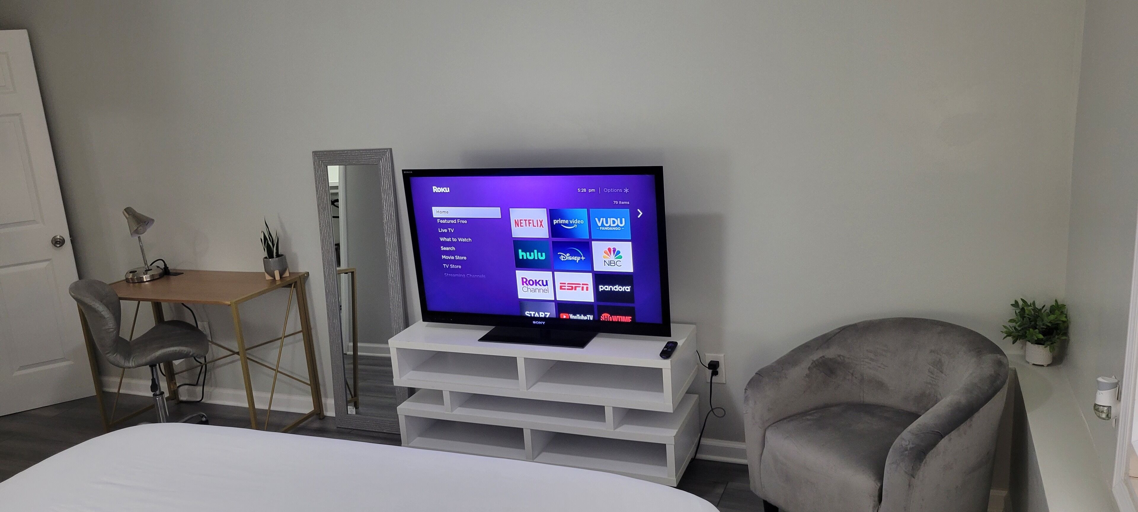 Una habitación moderna con una televisión de pantalla plana sobre un soporte blanco, un escritorio con lámpara, una silla y una cama.