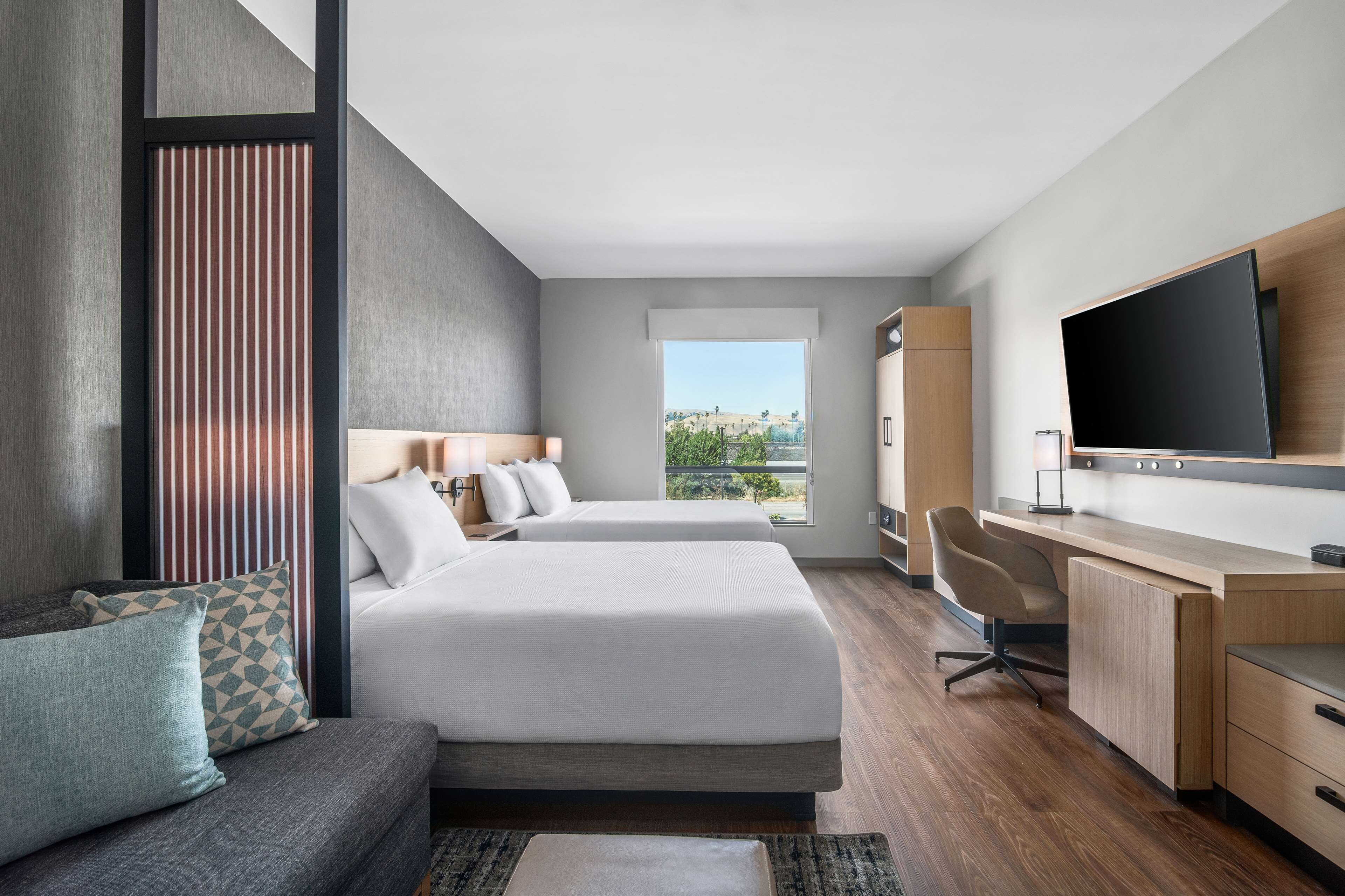 A modern hotel room with two beds, a desk, a TV, and a view of the outdoors.