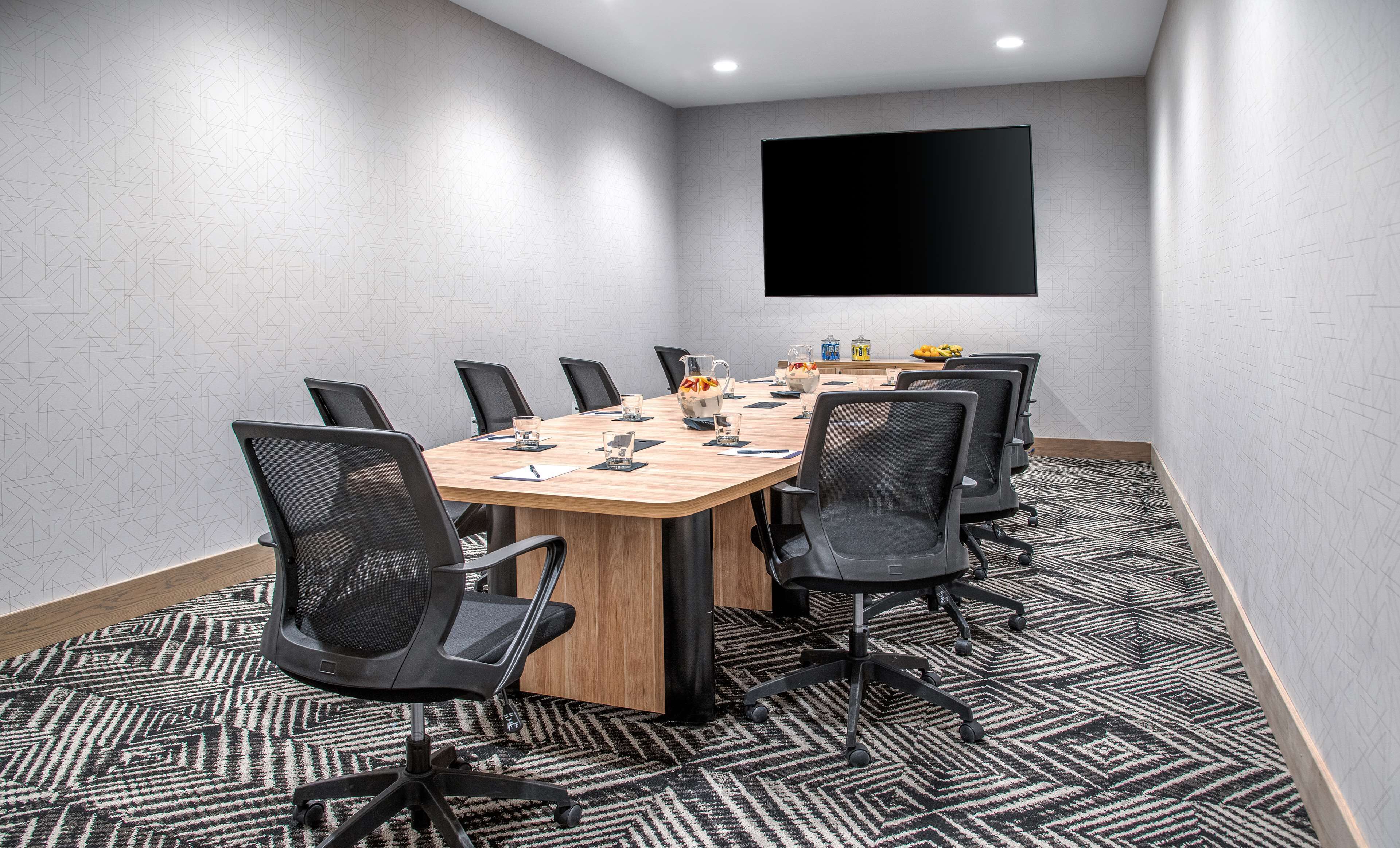 Uma sala de conferências com uma mesa comprida, cadeiras pretas, uma TV fixada na parede e um tapete estampado.