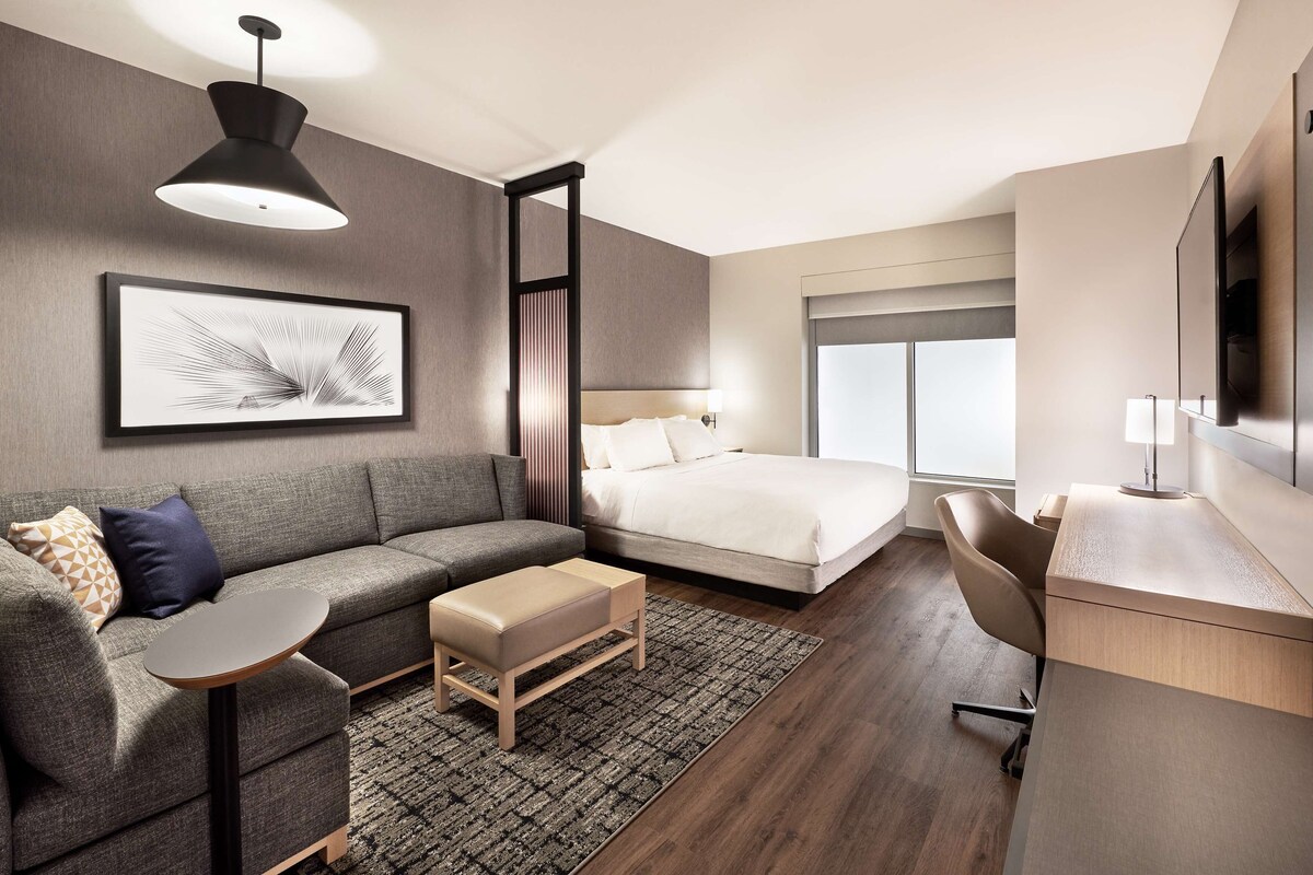A modern hotel room with a bed, sofa, desk, and a large window.