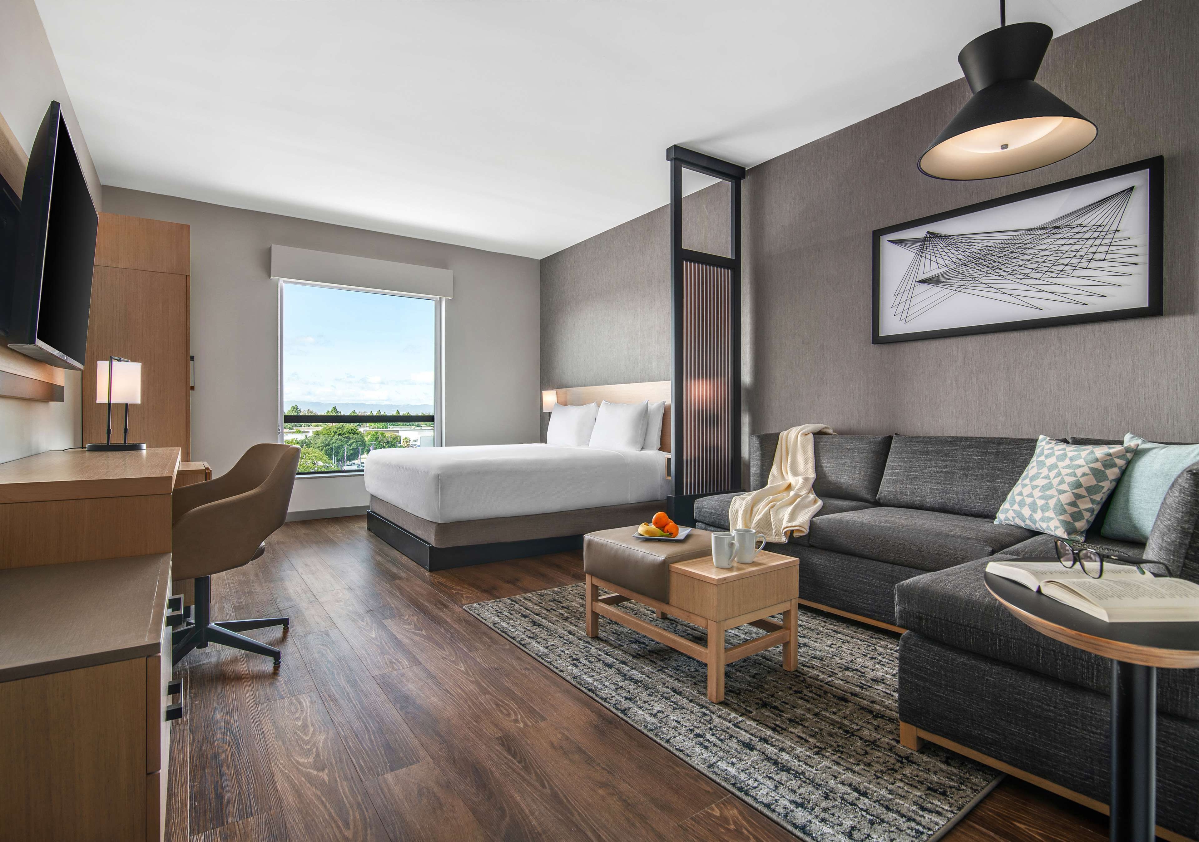 A modern hotel room with a bed, a sofa, a desk, and a large window offering a city view.