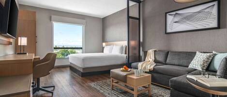 A modern hotel room with a bed, a sofa, a desk, and a large window offering a city view.