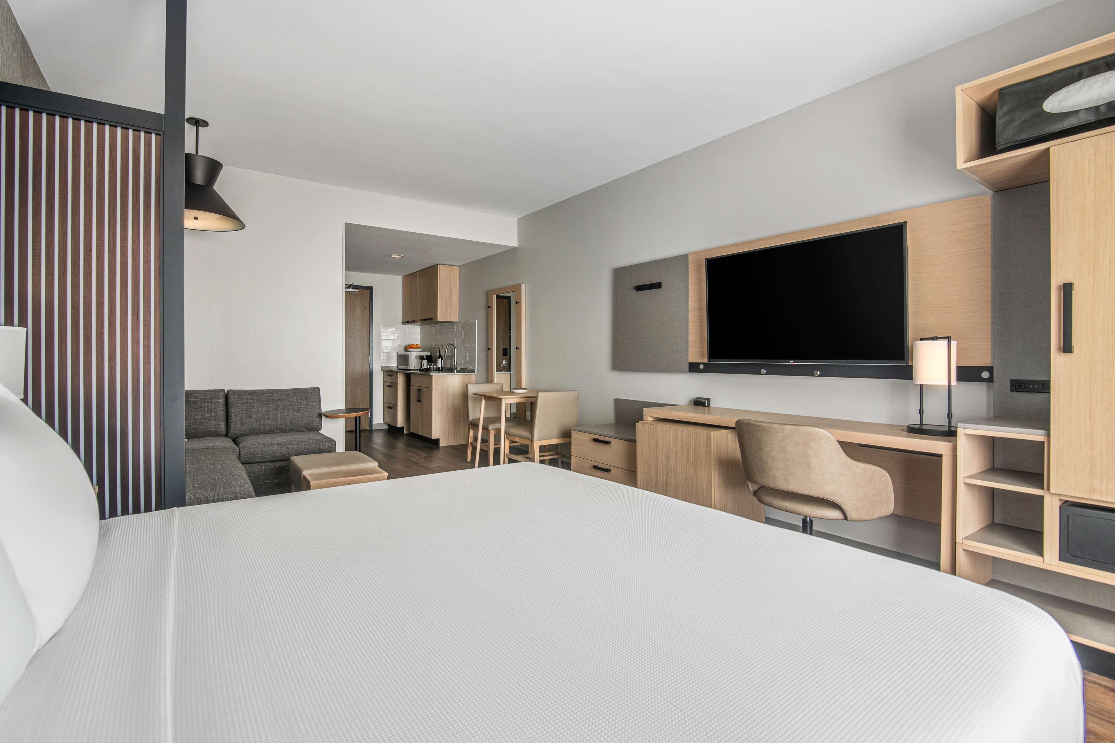A modern hotel room with a large bed, a desk, a TV, and a kitchenette.