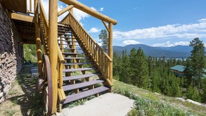 A wooden staircase leading to a building with a stone wall, surrounded by a mountainous landscape and trees.