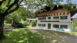 A traditional chalet with a sloped roof, wooden balconies, and a garden area with a bench and umbrella. - Wundervolle Erdgeschosswohnung mit Blick ins Grüne (Marquartstein)