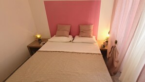 Een net opgemaakt bed met een roze hoofdbord, twee nachtkastjes met lampen, en een gordijn aan de rechterkant.