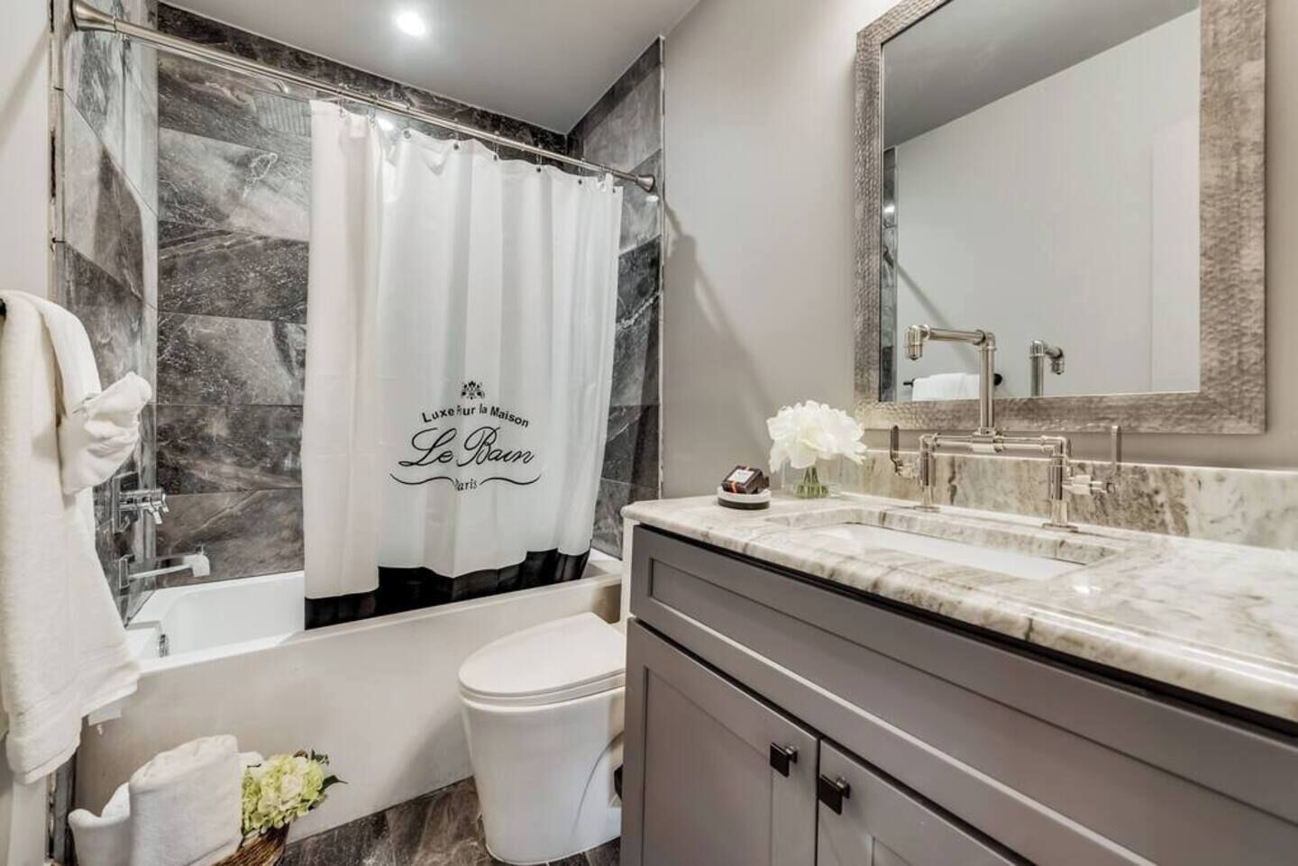 A modern bathroom with a bathtub, shower, and toilet. A white shower curtain with "Le Bain" text is visible.