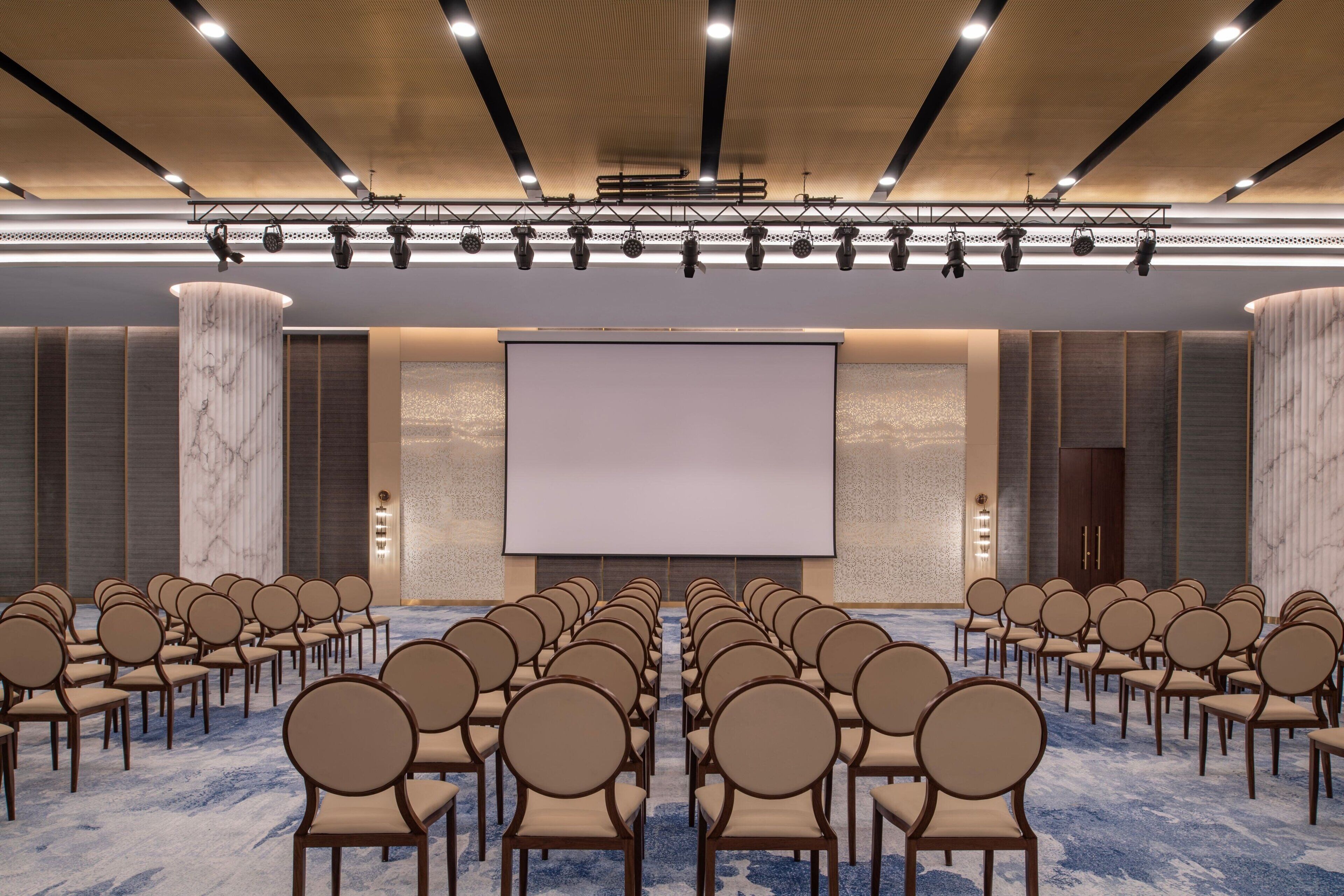 Un salón de conferencias con filas de sillas orientadas hacia una pantalla grande, un pilar de mármol y un diseño de techo moderno.