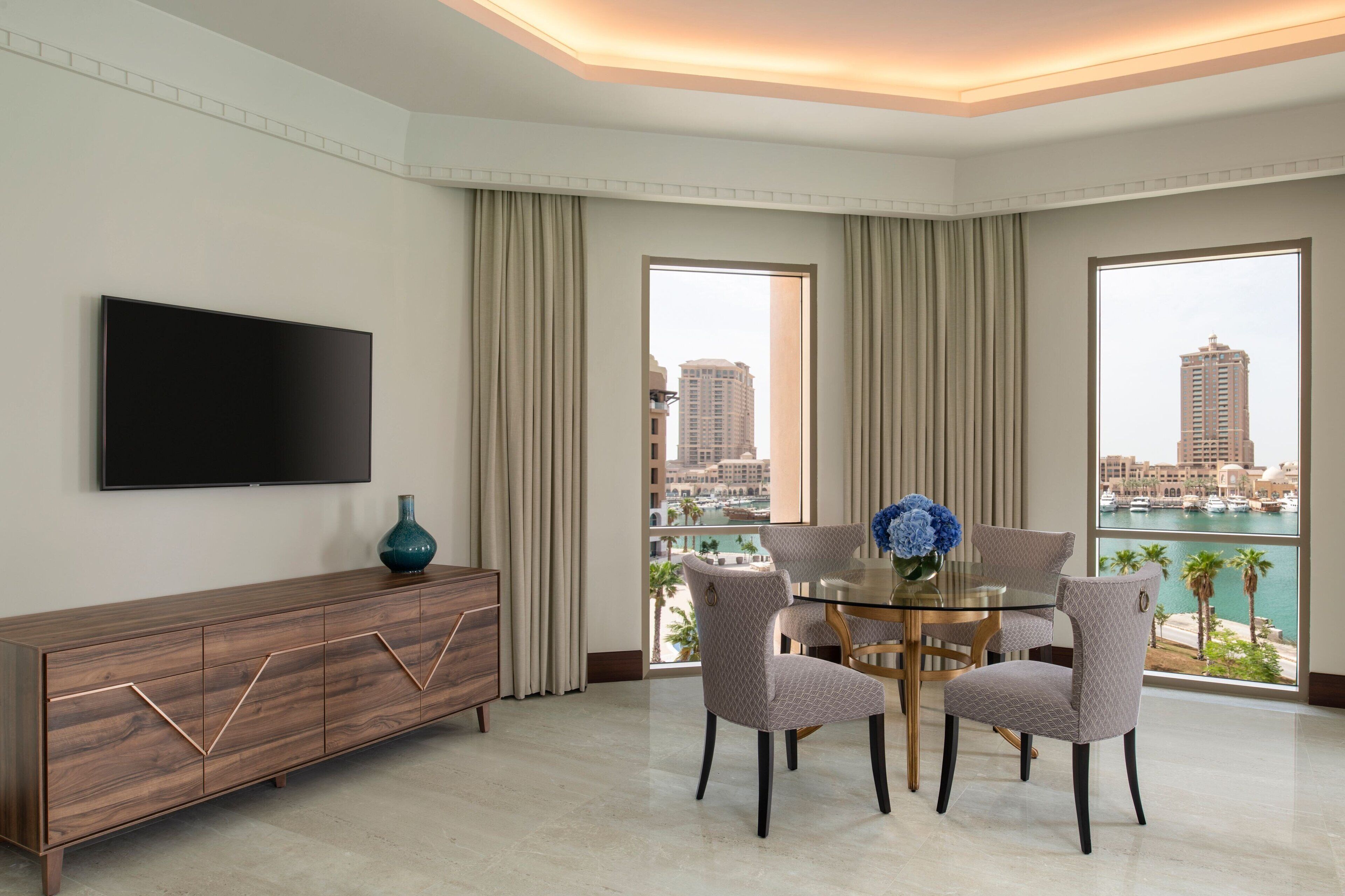 A modern hotel room with a large TV, a dining table with chairs, a view of the city, and a balcony with a view of the marina.