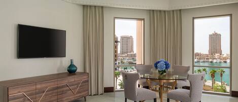 A modern hotel room with a large TV, a dining table with chairs, a view of the city, and a balcony with a view of the marina.