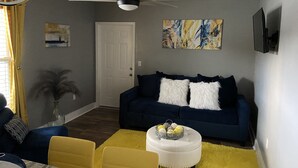A living room with a dark blue sofa, two white fluffy pillows, a white ottoman with a basket of decorative balls, and a yellow rug.