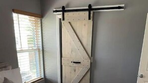 A bedroom with a bed, a sliding barn door, a window with blinds, and a bedside table with a remote control and towels.
