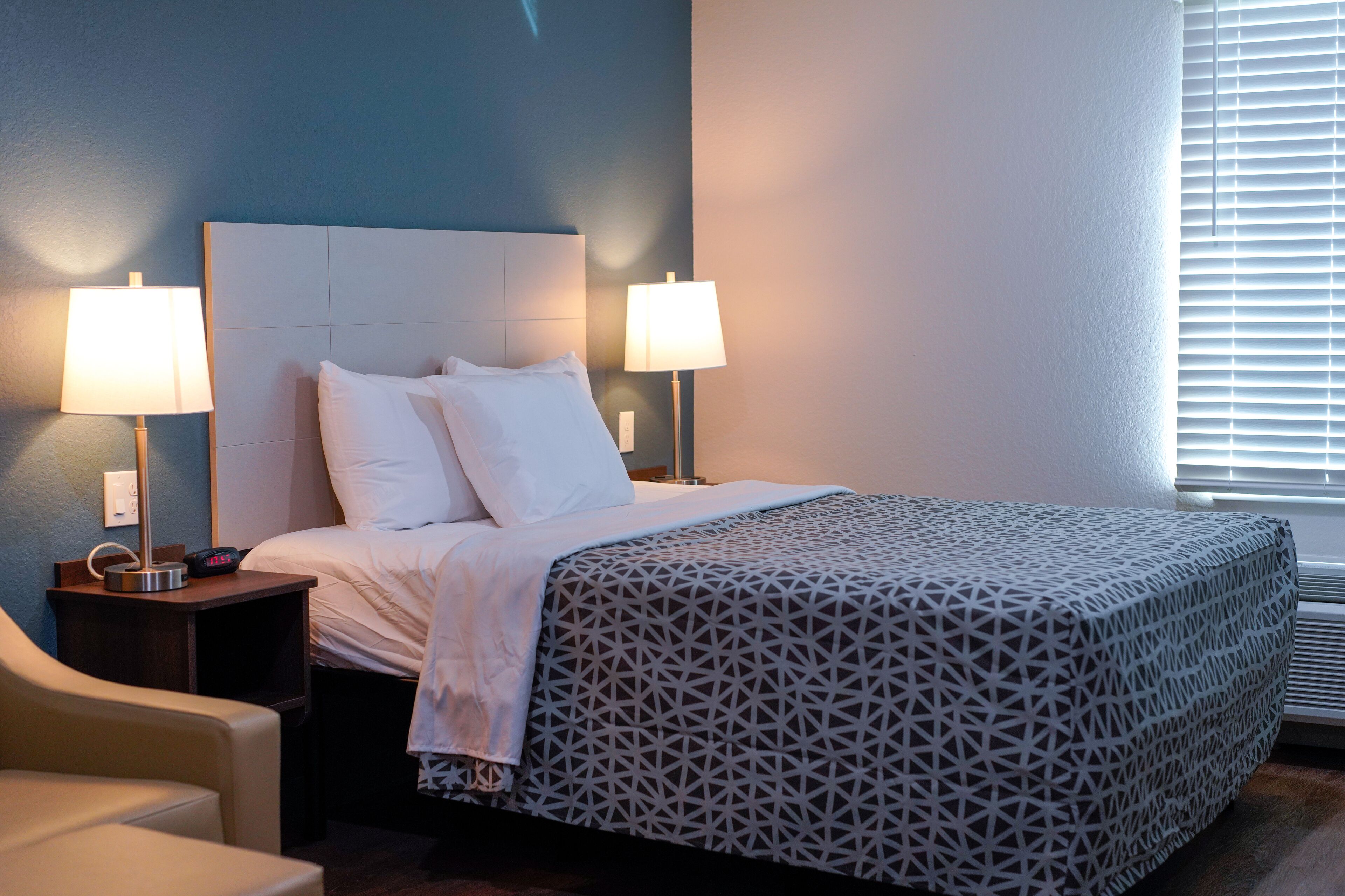 A hotel room with a bed, two bedside lamps, a chair, and a window with blinds.