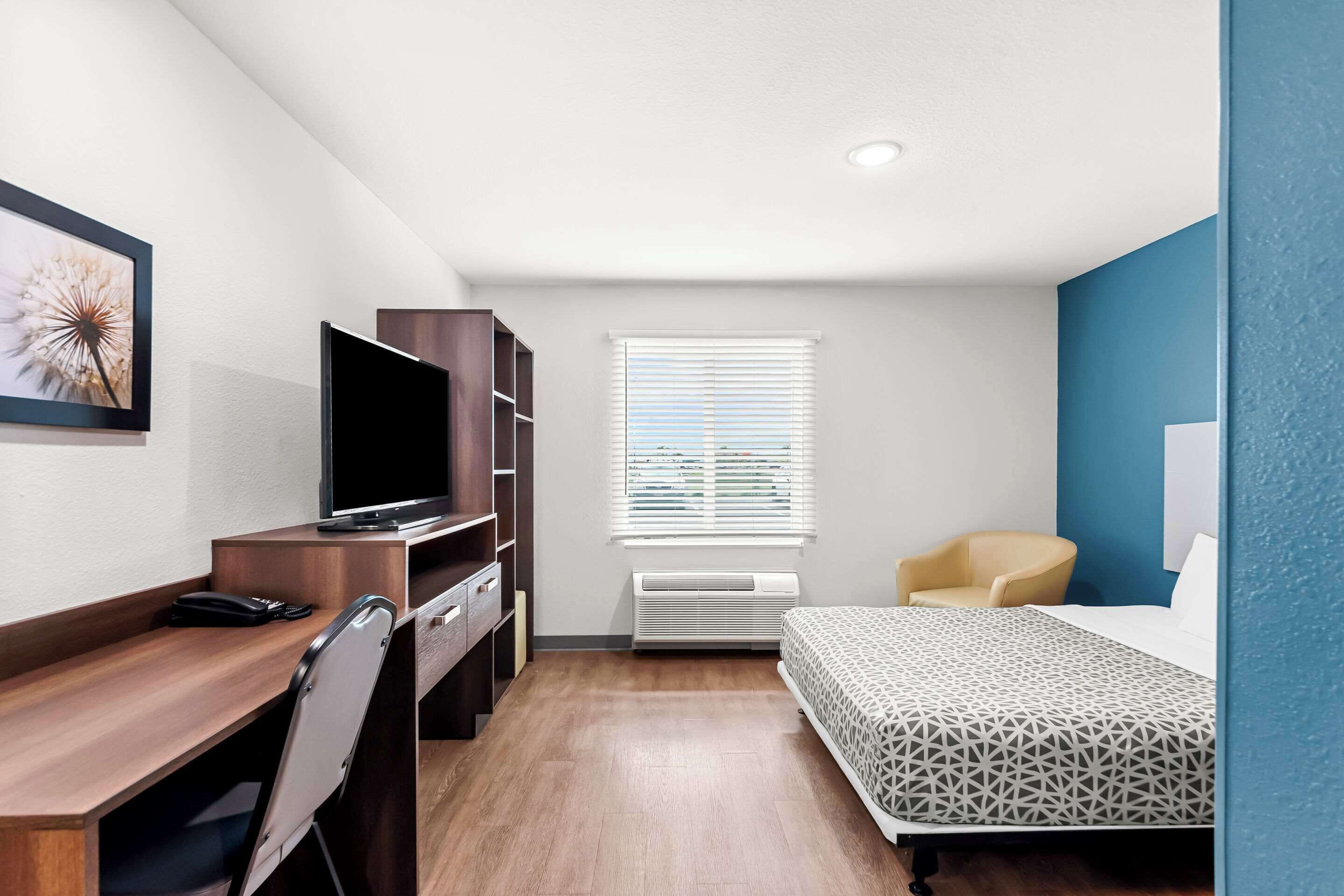 Habitación de hotel compacta con cama, escritorio con silla, televisor y ventana con persianas.