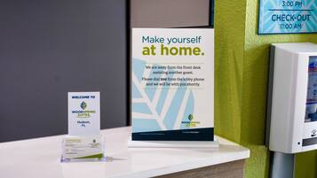 A hotel reception desk with a sign instructing guests to make themselves at home, a welcome sign, and a check-in/check-out schedule.