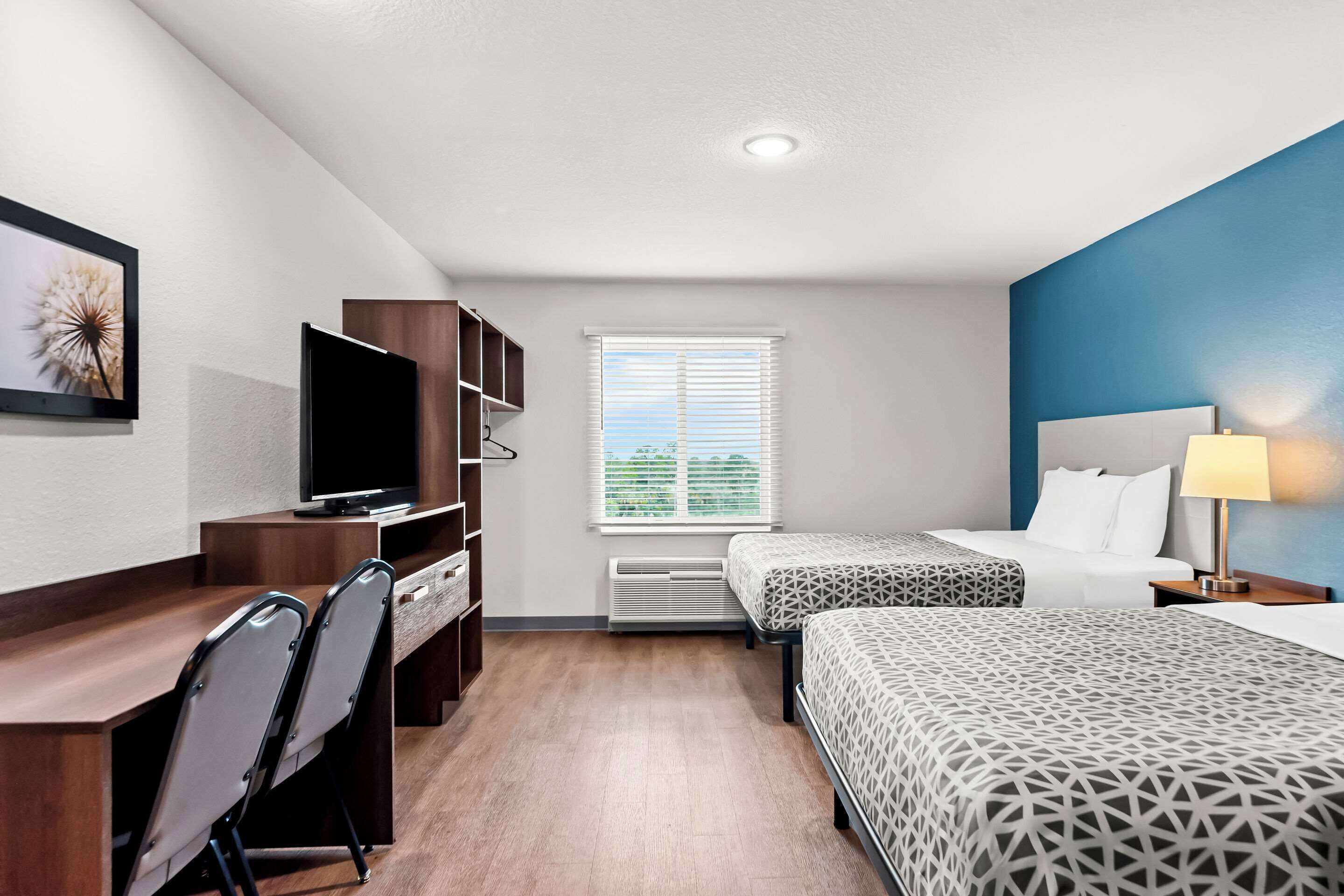 A hotel room with a desk, two beds, a TV, and a window with blinds.